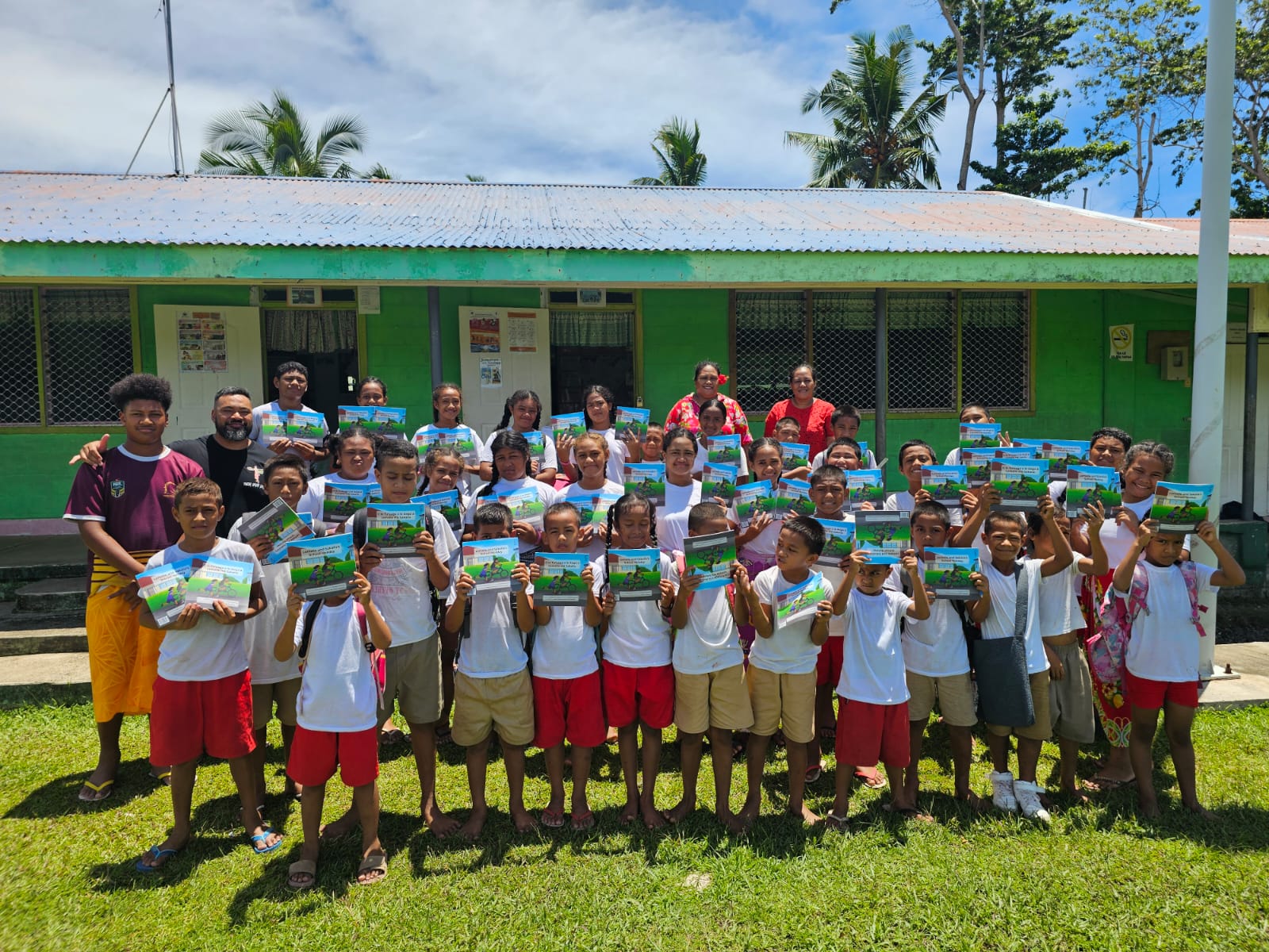 The Founder of Chief Book Series, Tony Laulu, poses with a group of primary school students in Savaii. Each person in the photo is holding up the book titled: Lemalie and Sakalia's School Holiday.