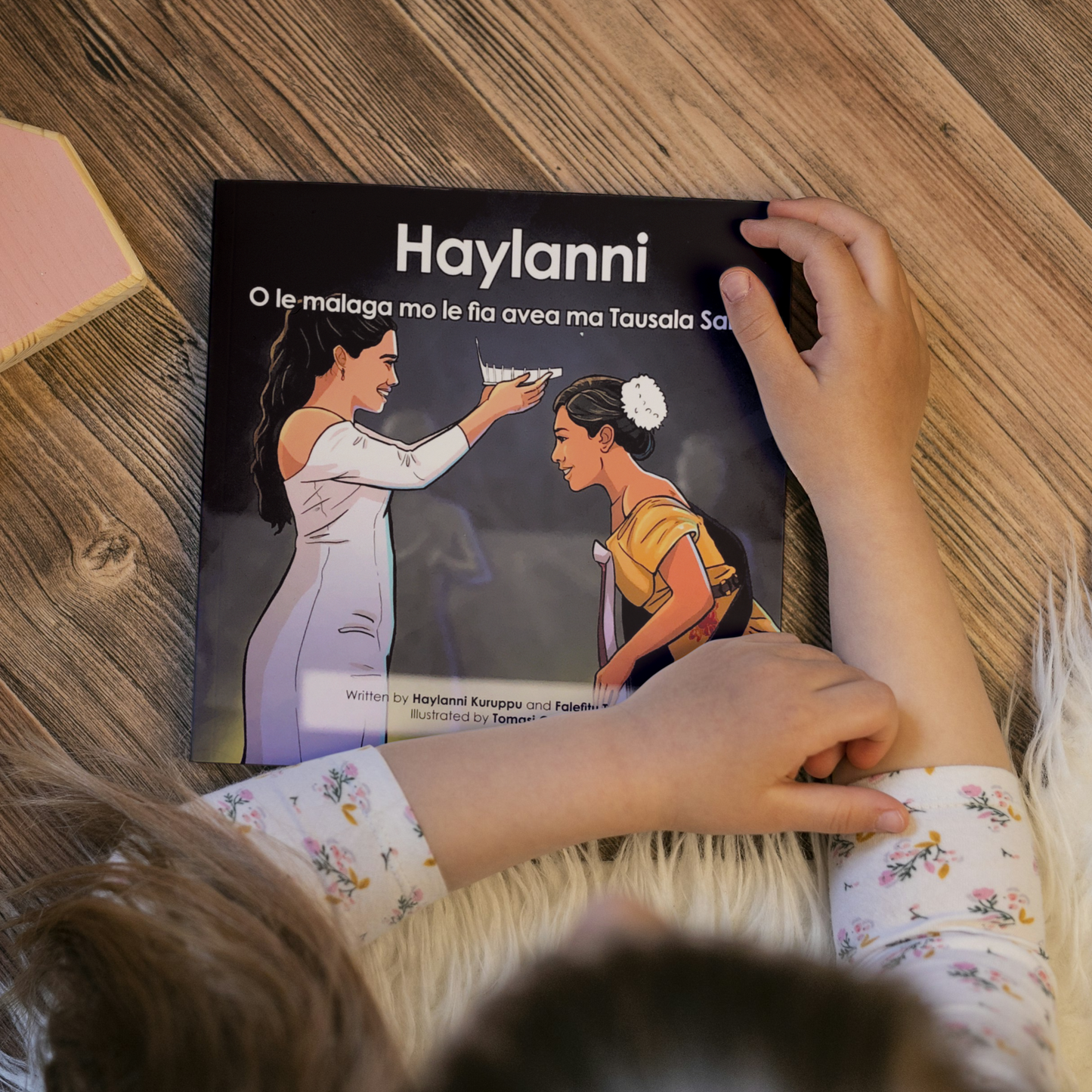 A young child reading a bilingual Samoan children's book titled "Lemalie and Sakalia’s School Holiday". The book is an illustration of two young boys riding their bikes during their school holiday.