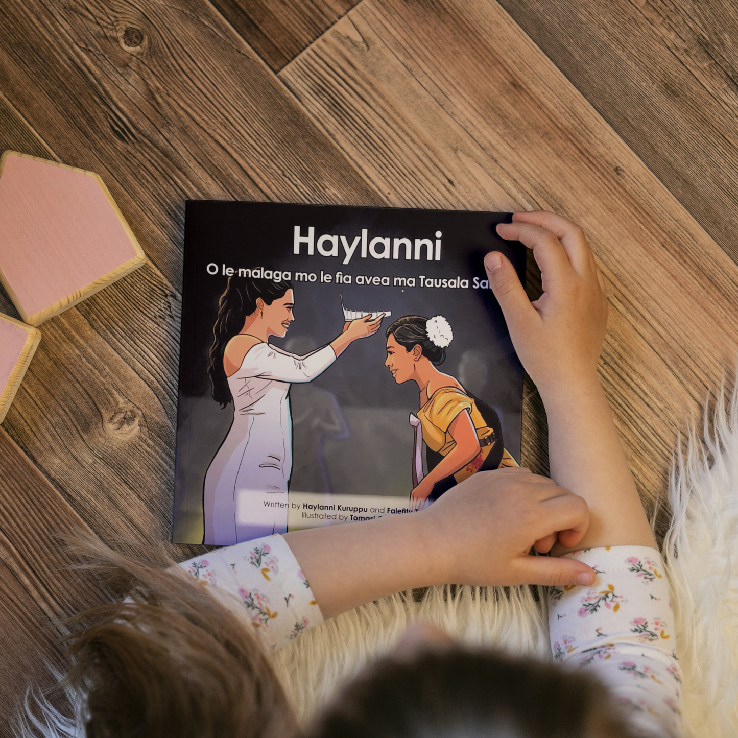 A young child reading a bilingual Samoan children's book titled "Haylanni: The Journey to Miss Samoa". The book is an illustration of a Fonoifafo Miss Samoa 2019-2021 crowning Haylanni as the new reigning Miss Samoa 2022-2023.