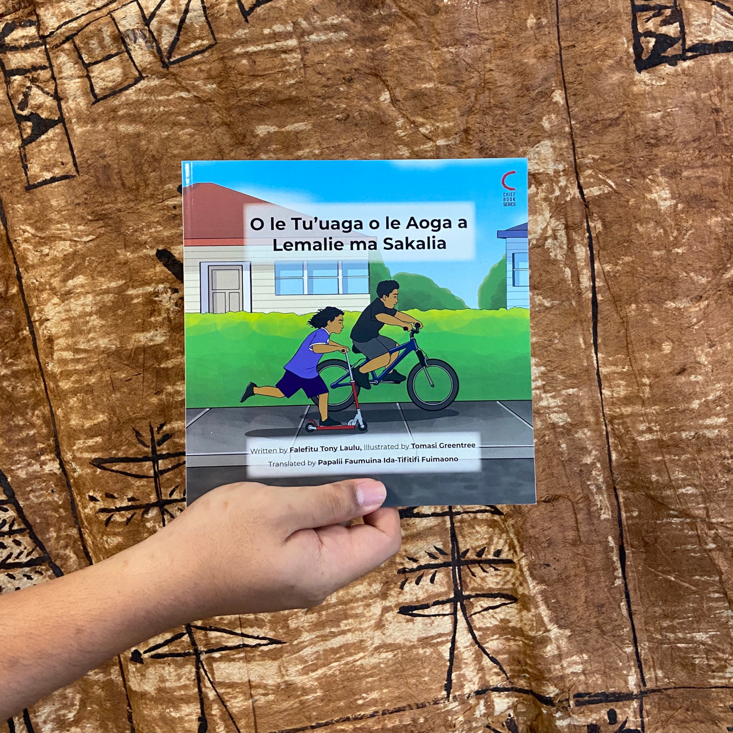 A hand model holding a bilingual Samoan children's book titled "Lemalie and Sakalia’s School Holiday". The book is an illustration of two young boys riding their bikes during their school holiday.