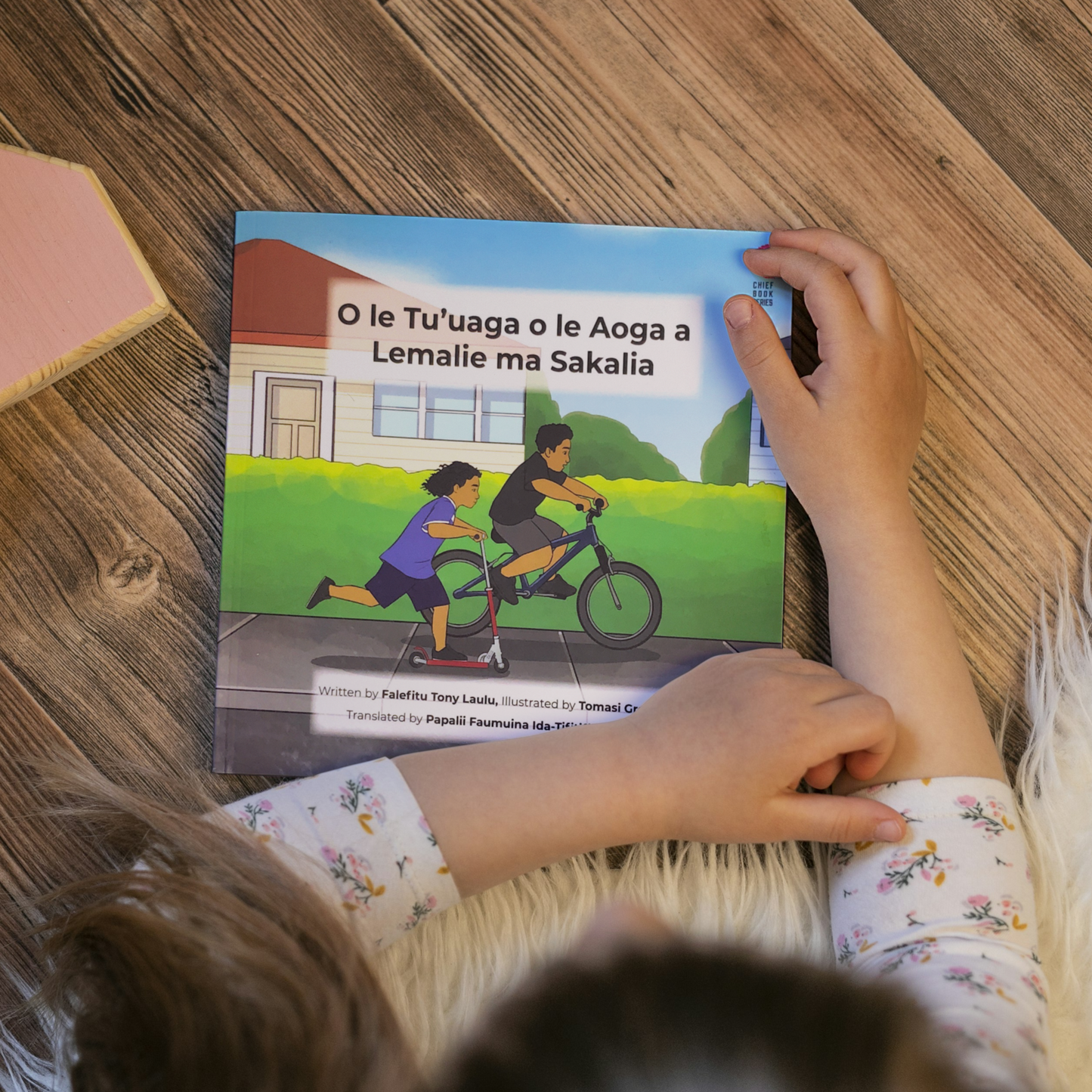 A young child reading a bilingual Samoan children's book titled "Lemalie and Sakalia’s School Holiday". The book is an illustration of two young boys riding their bikes during their school holiday.