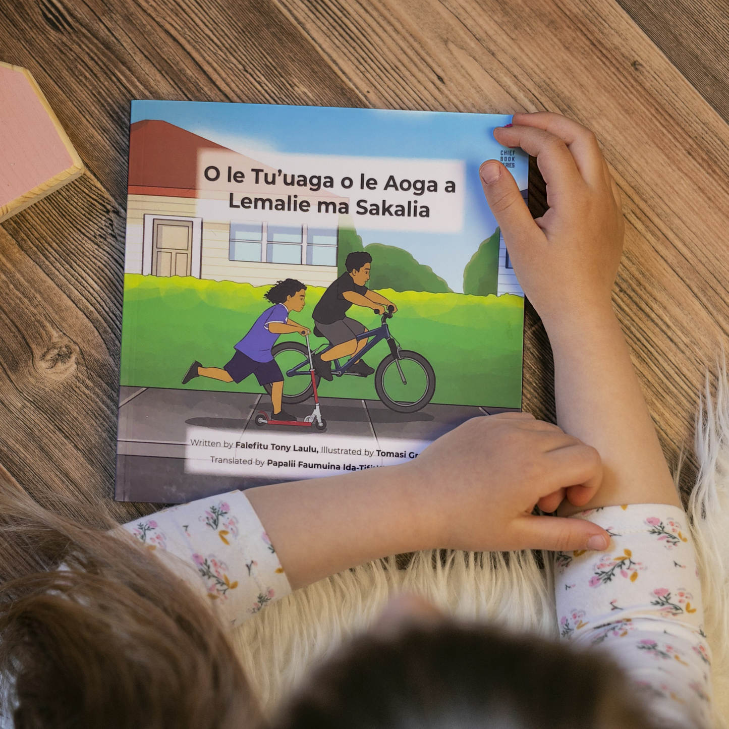 A young child reading a bilingual Samoan children's book titled "Lemalie and Sakalia’s School Holiday". The book is an illustration of two young boys riding their bikes during their school holiday.