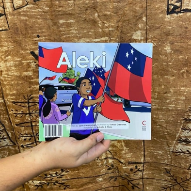 A hand model holding a bilingual Samoan children's book titled "Aleki". The book is an illustration of a young boy holding the Samoan Flag proudly during a parade.
