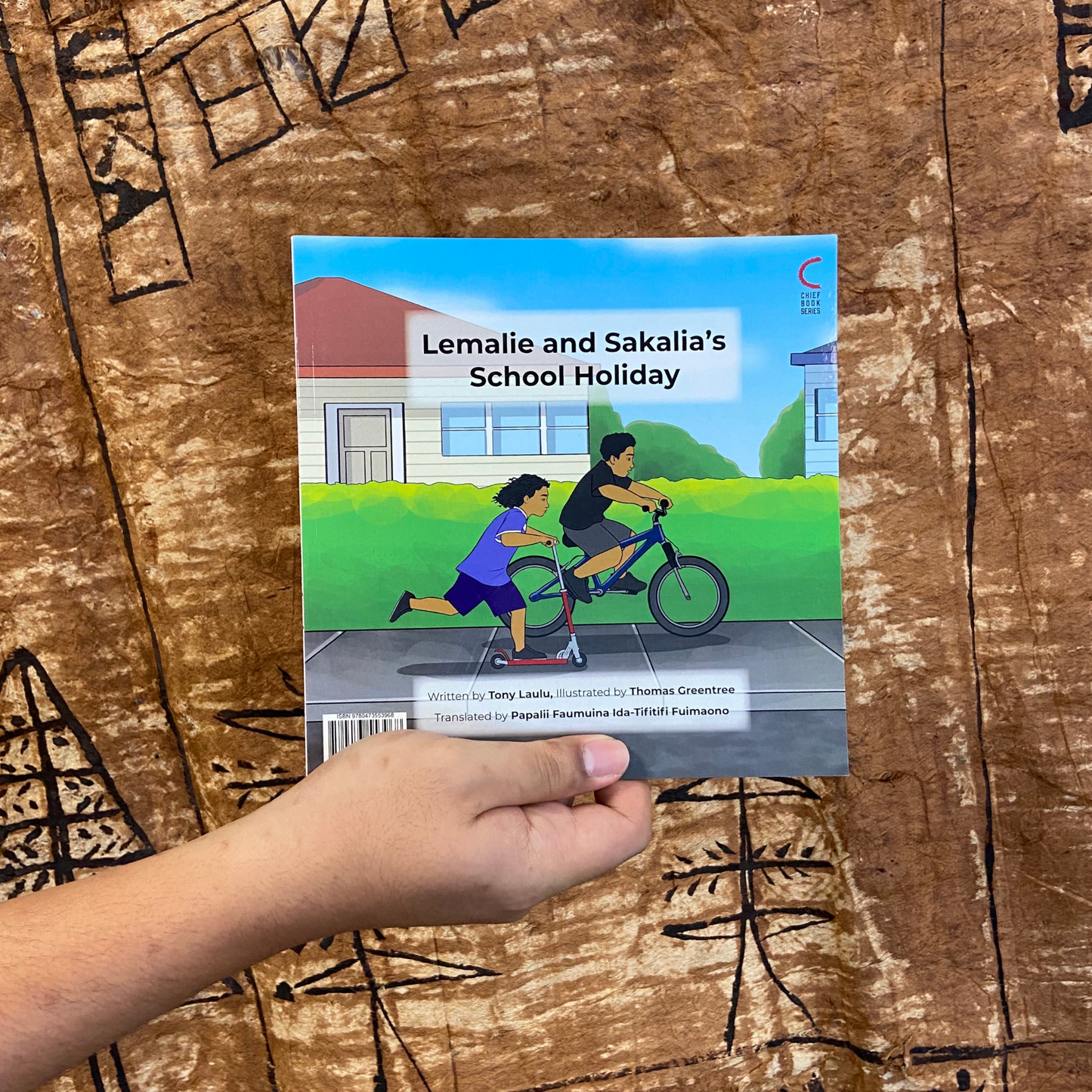A hand model holding a bilingual Samoan children's book titled "Lemalie and Sakalia’s School Holiday". The book is an illustration of two young boys riding their bikes during their school holiday.