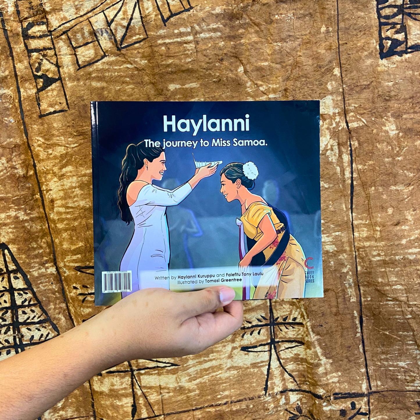 A hand model holding a bilingual Samoan children's book titled "Haylanni: The Journey to Miss Samoa". The book is an illustration of a Fonoifafo Miss Samoa 2019-2021 crowning Haylanni as the new reigning Miss Samoa 2022-2023.