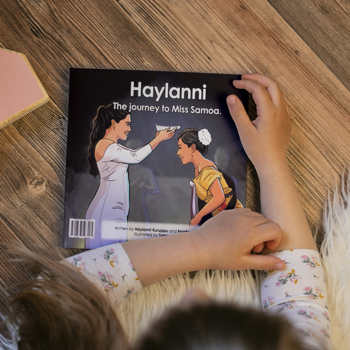 A young child reading a bilingual Samoan children's book titled "Haylanni: The Journey to Miss Samoa". The book is an illustration of a Fonoifafo Miss Samoa 2019-2021 crowning Haylanni as the new reigning Miss Samoa 2022-2023.