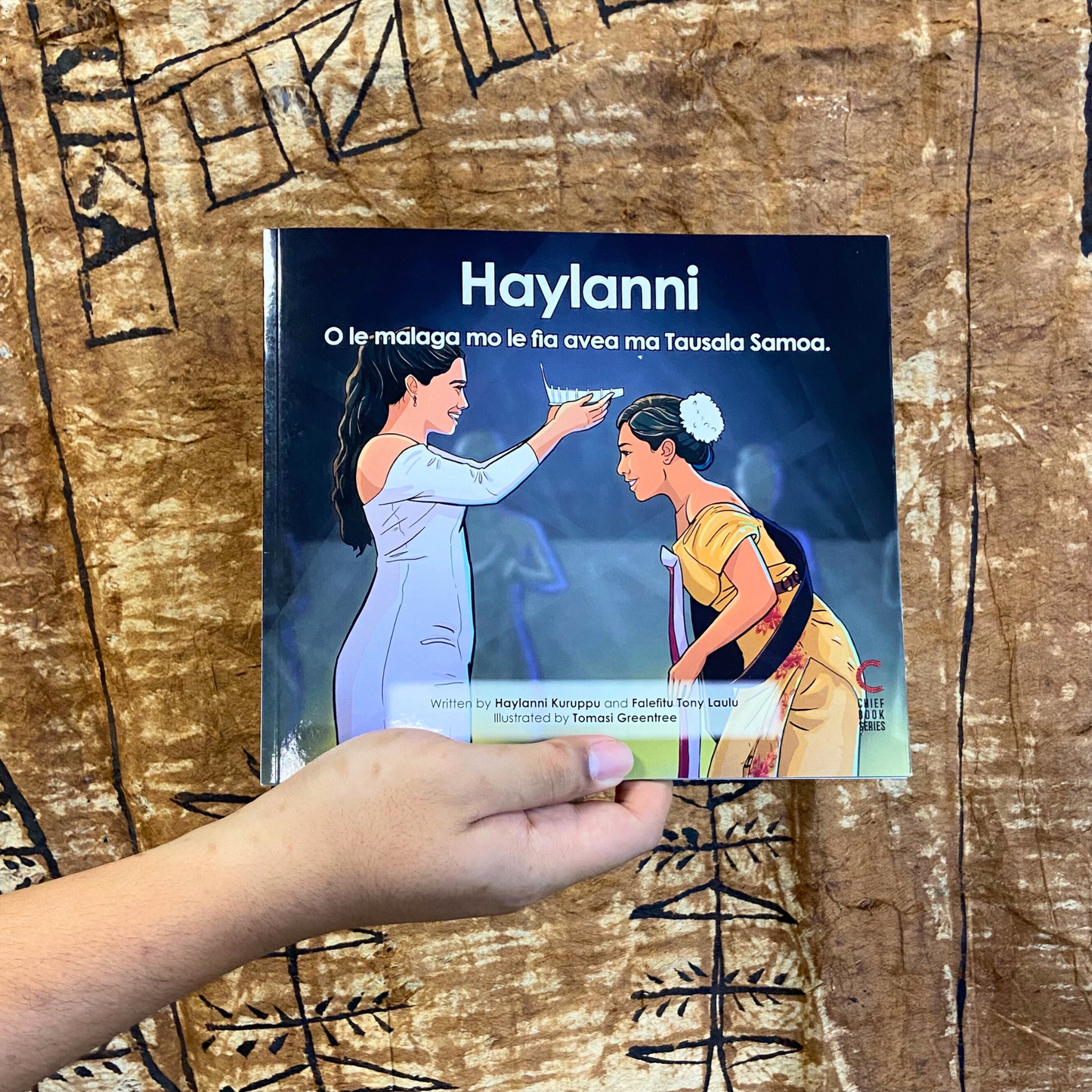 A hand model holding a bilingual Samoan children's book titled "Haylanni: The Journey to Miss Samoa". The book is an illustration of a Fonoifafo Miss Samoa 2019-2021 crowning Haylanni as the new reigning Miss Samoa 2022-2023.