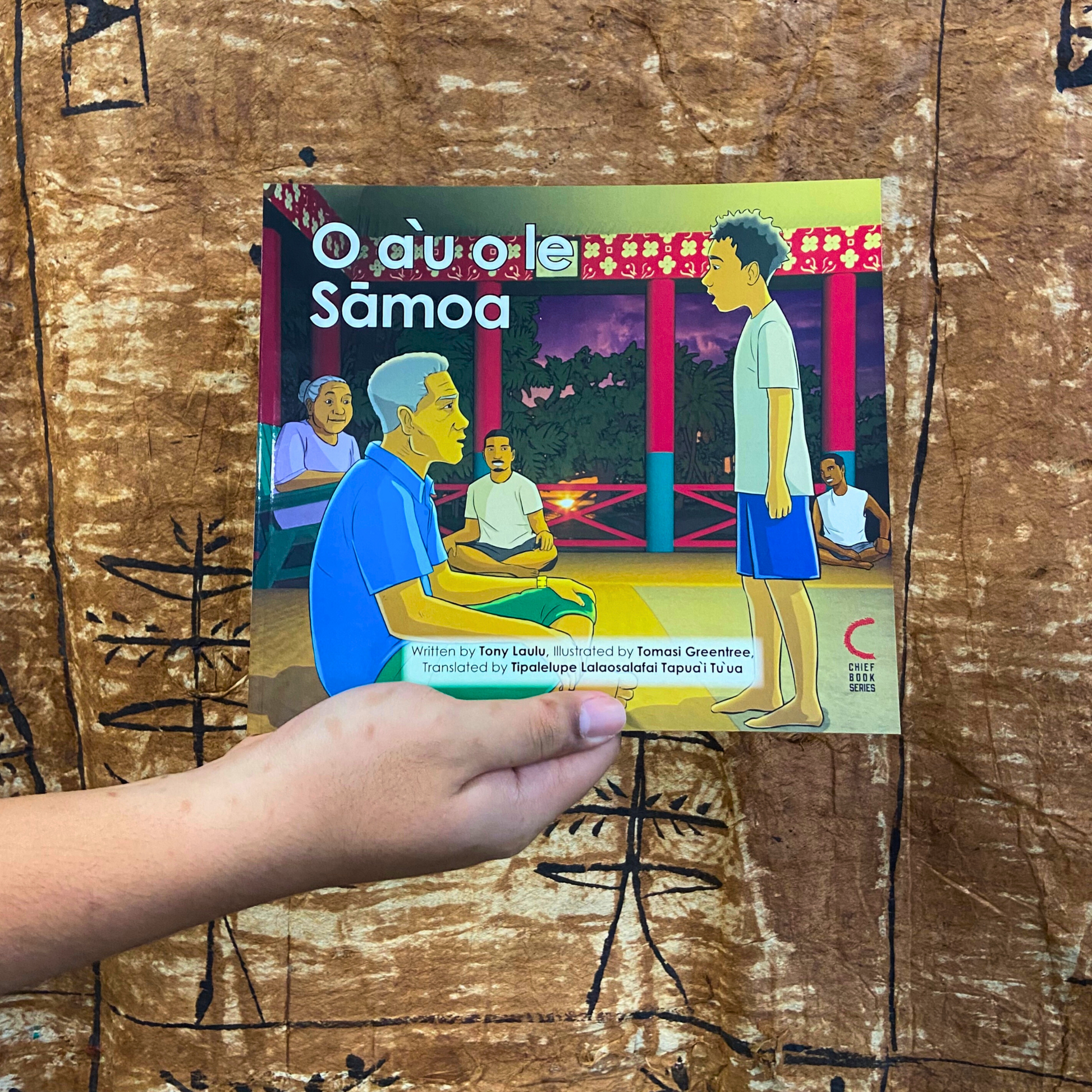 A hand model holding a bilingual Samoan children's book titled "O a’u o le Samoa". The book is an illustration of a young Samoan boy standing directly in front of an elderly man in a Fale Samoa.