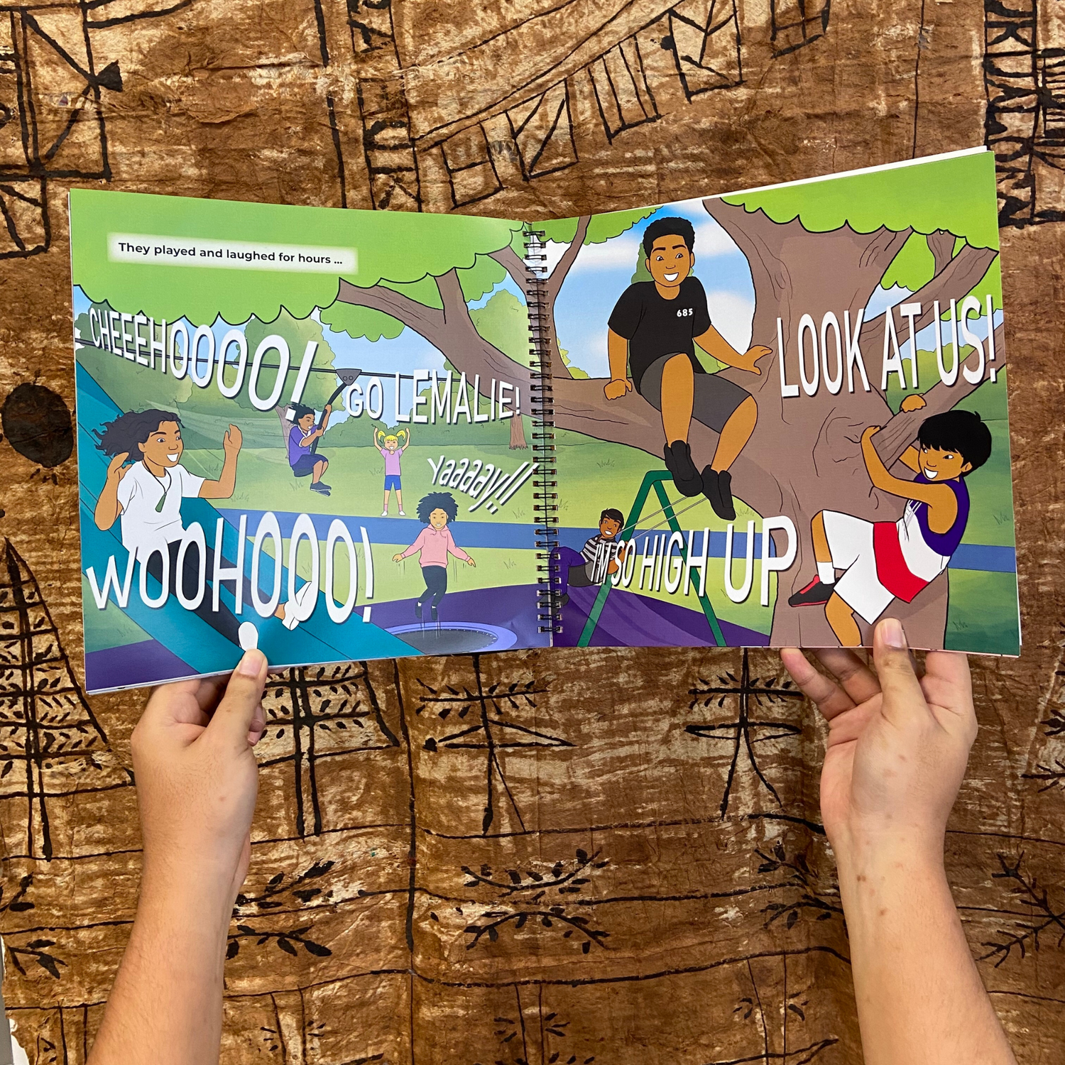 A hand model holding a bilingual Samoan children's book titled "Lemalie and Sakalia’s School Holiday". The book is an illustration of two young boys riding their bikes during their school holiday.