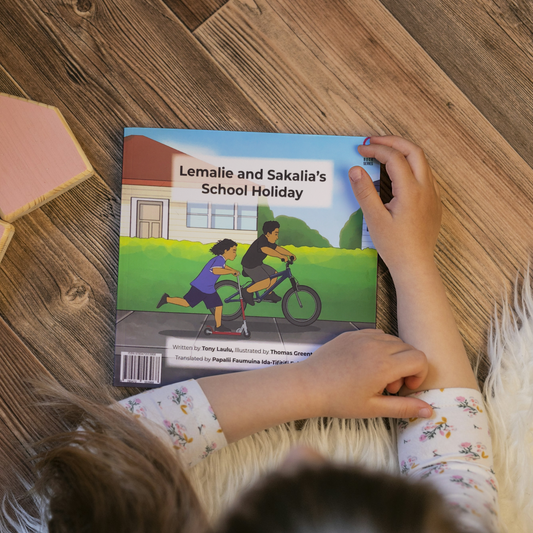 A young child reading a bilingual Samoan children's book titled "Lemalie and Sakalia’s School Holiday". The book is an illustration of two young boys riding their bikes during their school holiday.