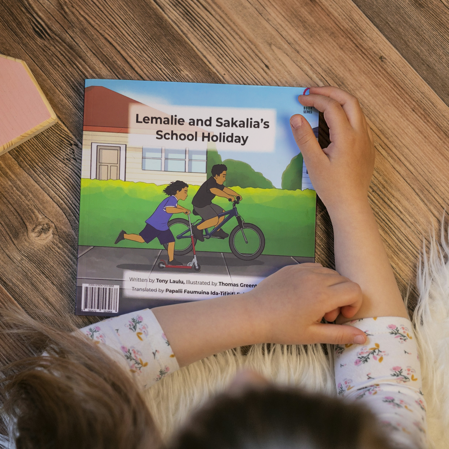 A young child reading a bilingual Samoan children's book titled "Lemalie and Sakalia’s School Holiday". The book is an illustration of two young boys riding their bikes during their school holiday.