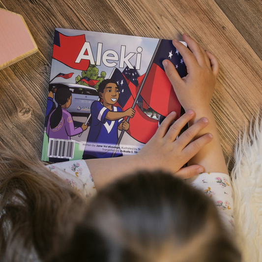 A young child reading a bilingual Samoan children's book titled "Aleki". The book is an illustration of a young boy holding the Samoan Flag proudly during a parade.