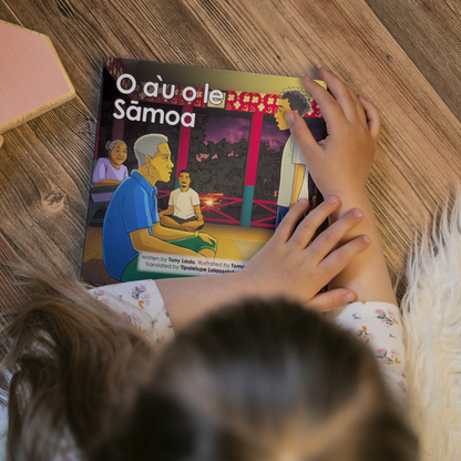 A young child reading a bilingual Samoan children's book titled "O a’u o le Samoa". The book is an illustration of a young Samoan boy standing directly in front of an elderly man in a Fale Samoa.